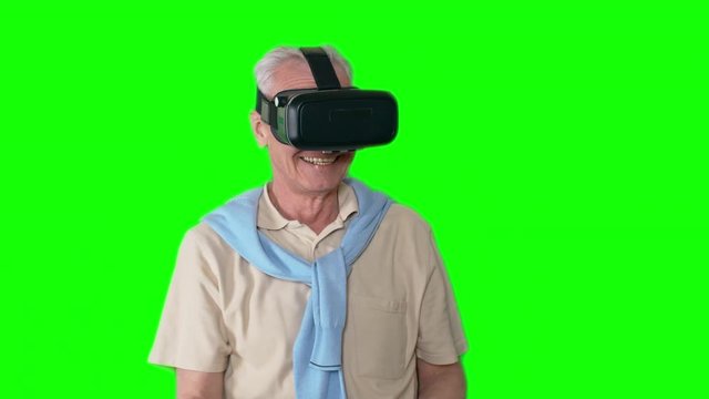 Widely smiling old man with grey hair standing against green screen background wearing virtual reality goggles and waving his arms around