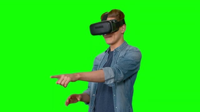 Young man in virtual reality headset standing against green background looking around and gesturing