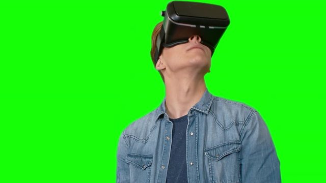 Serious young man in virtual reality headset standing against green screen background and looking around, then taking it off