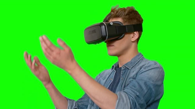 Young man in virtual reality headset standing against green screen catching something invisible