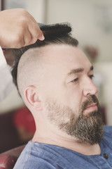 Male barber makes hair styling using a hair dryer of a adult bearded man with a mohawk