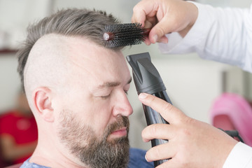 Male barber makes hair styling using a hair dryer of a adult bearded man with a mohawk