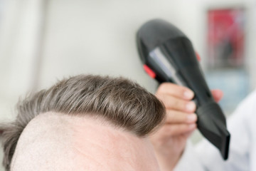 Male barber makes hair styling using a hair dryer of a adult man with a mohawk