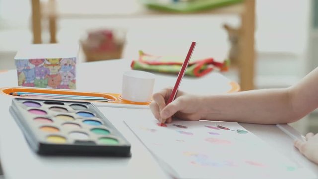 Color red pencil in child hand. Paints and drawing on the table. Child draws with colored pencils. School desk. Children's drawing. The child is busy drawing.