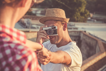 Young trendy guy taking photos of his girlfriend with retro vintage camera and holding her hand in nature. Travel, vacation and love concepts. 