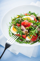 Fresh salad with tuna, tomatoes, eggs, arugula and mustard on blue wooden background close up. Healthy food.