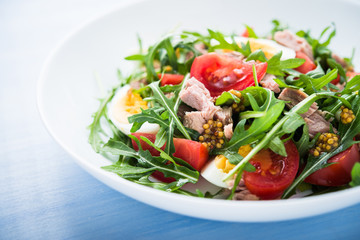 Fresh salad with tuna, tomatoes, eggs, arugula and mustard on blue wooden background close up. Healthy food.