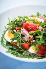 Fresh salad with tuna, tomatoes, eggs, arugula and mustard on blue wooden background close up. Healthy food.