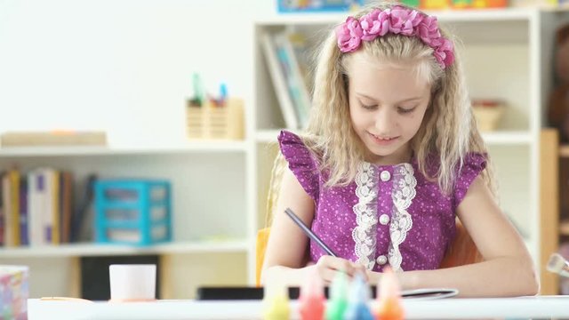 Beautiful little girl in a lavender dress draws. The girl smiles. Leisure children. Child development. Girl draws. Children's creativity.