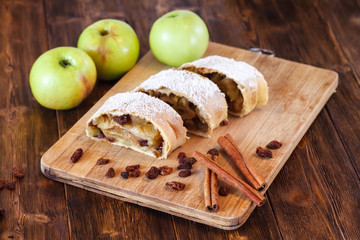 Apple strudel with icing sugar, cinnamon sticks, wooden background