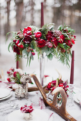 Wedding table setting in marsala colors with plates, cutlery, red floral compositions, candles, velvet napkins, deer horns on table covered with snow