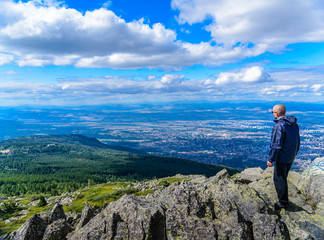 Middle aged man in sports clothing and sunglasses on top of a mountain, overlooking the city of Sofia, Bulgaria from high altitude - beautiful landscape with city shapes in the far background