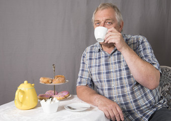 Mature man drinking a cup of tea whilst sitting down at a table. Taken on a grey background. 