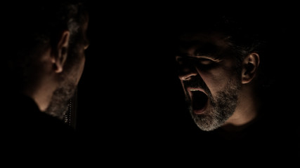 Fototapeta premium Angry man shouting in front of mirror at his reflection in a dark room