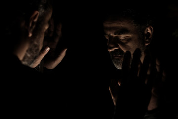 Man in despair with raised hands and bowed head, in a low light room looking in front of mirror