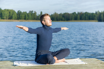 Young man relaxing and enjoying a first sun rays and freedom in the morning. Man practicing yoga on the lake footbridge.