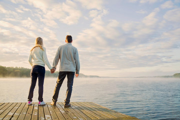 Fototapeta premium Young couple staring at beautiful, peaceful landscape with sky and lake in the mist on the footbridge in autumn early morning.