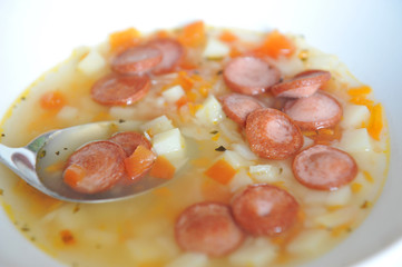 Vegetable soup with added pieces of frankfurter sausages in a white plate.