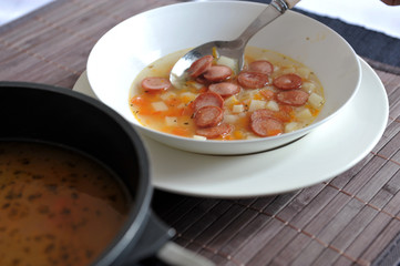Vegetable soup with added pieces of frankfurter sausages in a white plate.