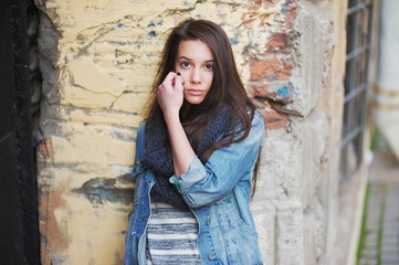Sad young brown-eyed girl with long hair in a black scarf, and  denim jacket against the background of an old brick wall, close-up.