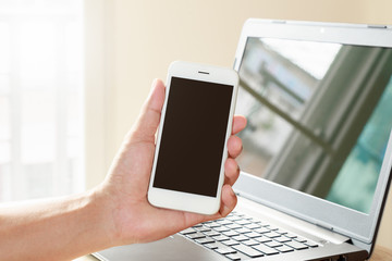 Young man hand holding smartphone black screen with laptop on desk