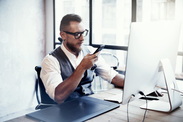 Bearded Stylish Young Man Wearing Glasses White Shirt Waistcoat Working Modern Loft Startup Process.Creative Person Using Smartphone Texting Message.Drawing Tablet Desktop Computer Wood Table.Blurred.