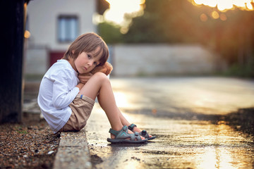 Sad little boy, sitting on the street in the rain, hugging his t