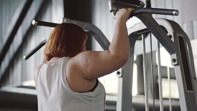 Tilt up of fat woman hanging on bar and doing pull-up exercises in the gym