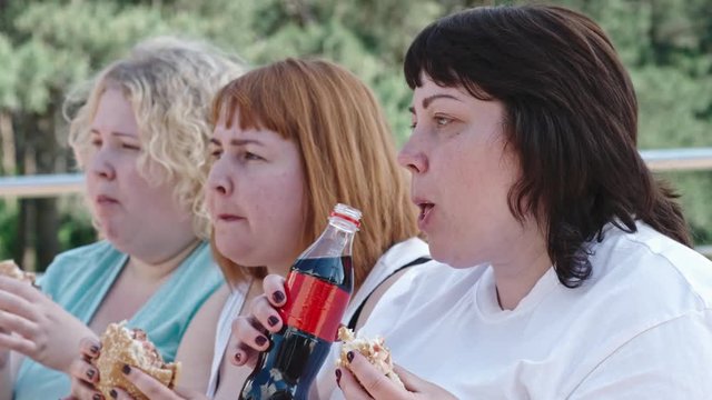 Closeup of three overweight women eating burgers and one of them drinking coke from plastic bottle