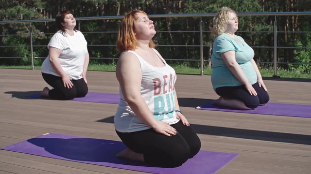 Three overweight woman sitting with closed eyes outdoor on mats while doing yoga exercise