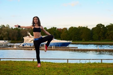 Fitness sport woman doing streching with legs during outdoor cross training workout. Girl making evening exercises. Female in a black tank top and tights under bright sun.