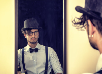 Portrait of brunette young man in glasses, hat, bow-tie, suspenders and shirt looking at himself in a mirror. Vintage look