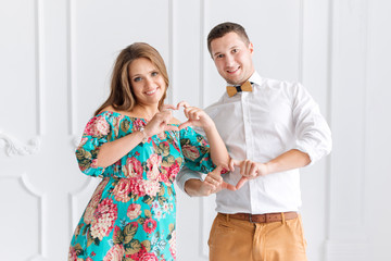 Beautiful pregnant couple happy together expecting a child. Man and woman in white minimalistic interior showing hearts with hands. Focus on fingers. Share the love concept.