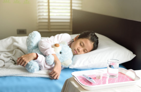 Thermometer and pills medicine in glass on table in front of out of focus sleeping child for health and illness concept, young Asian girl hugs the blue teddy bear.