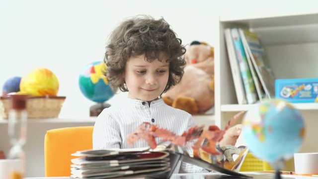 Child and book. Cute boy with black curly hair opens the book of fairy tales. Boy with black curly hair look a book with pictures. Curly boy in the white shirt. Globe on the table. Child's room.