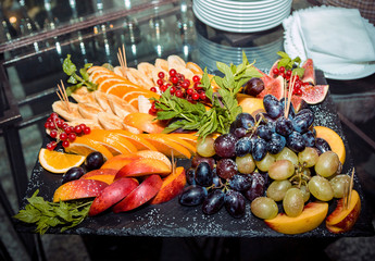 Fresh ripe fruits, berries and mint arrangement on a black stone tray. Sliced peach, orange, banana, fig, grape and viburnum clusters, decorated by mint and sugar powder