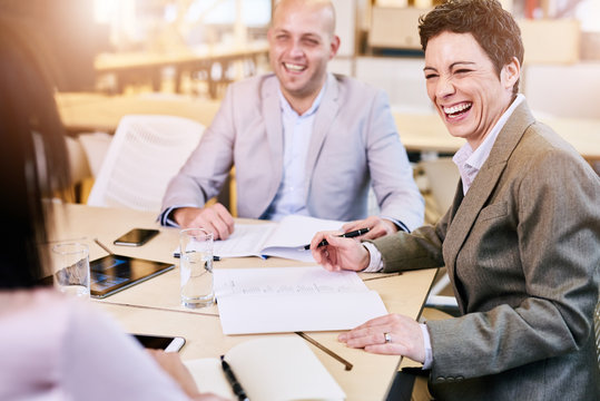 over the shoulder image of business executives laughing happily during a meeting seated at a conference table looking toward the camera.