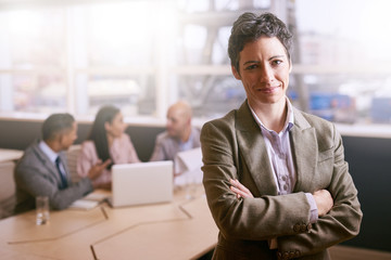 Confident businesswoman smiling at the camera while standing with her arms crossed in front of her colleagues that are seated at the table behind her.