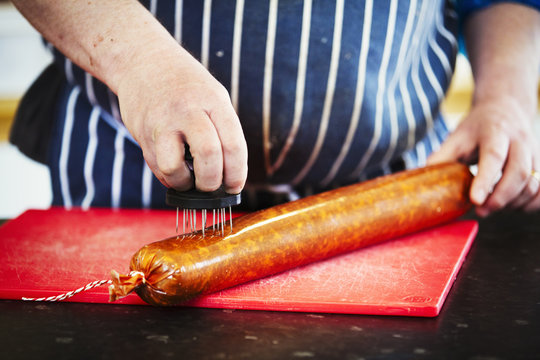 Close up of a butcher wearing a striped blue apron, pricking holes into the casing of a Chorizo sausage.