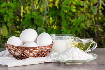 Пшеничная мука, куриные яйца, подсолнечное масло и чашка молока на деревянном столе