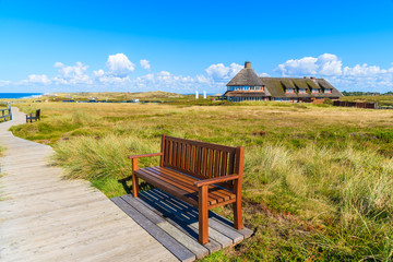 Bench on walking path along a coast of Sylt island and typical Frisian guest house in background,...