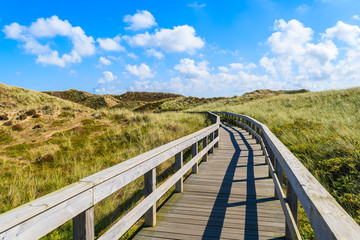 Fototapeta na wymiar Wooden walkway to beach among sand dunes and sunny blue sky with white clouds, Sylt island, Germany