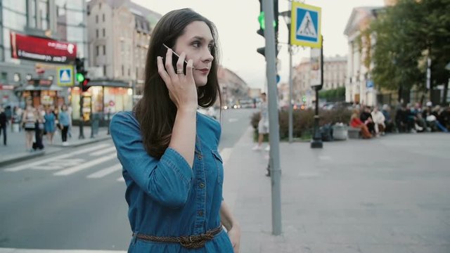 Smiling pretty woman in a blue denim dress talks on the phone in the centre of busy street. Slow mo