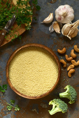 Raw couscous in wooden bowl, parsley, broccoli, cashew nuts and garlic on the side, photographed overhead on slate with natural light