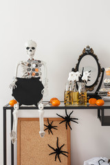 A table decorated for Halloween. A skeleton holding a black cauldron shaped chalkboard. Glass bottles with apple juice and paper straws with marshmallow. A decorative mirror and clementines. 