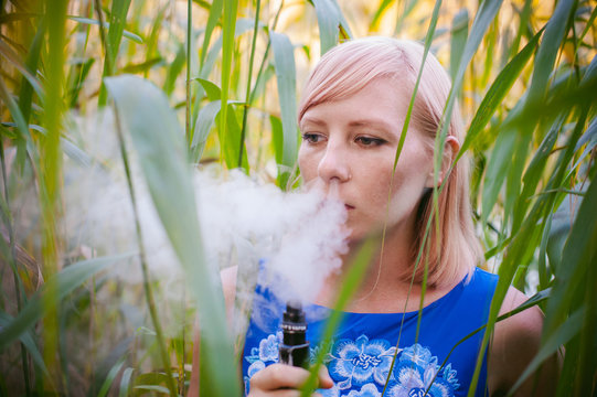 portrait nude vaping girl in sedge. young blonde woman standing in the reeds and smoking an electronic cigarette, blowing the smoke vapor