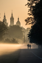 Fototapeta Unidentified men silhouettes in the mist, Blonia meadow in Krakow, Poland, with St Mary's church and Town Hall towers in the background. obraz
