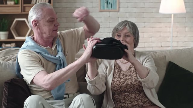 Elderly man sitting beside smiling senior woman and helping her to put virtual reality headset on