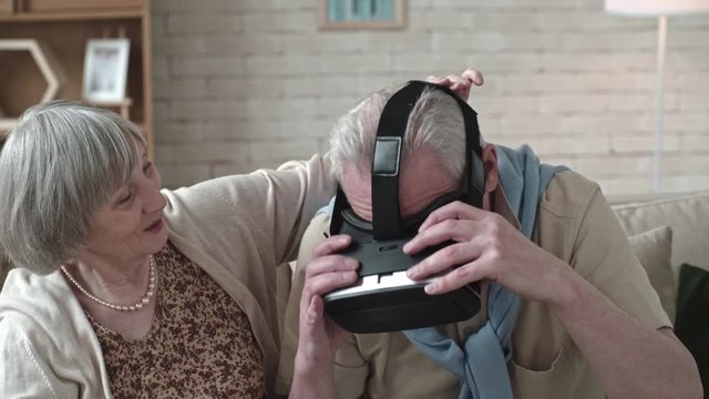 Elderly woman helping senior man to put on virtual reality goggles, then discussing experience 