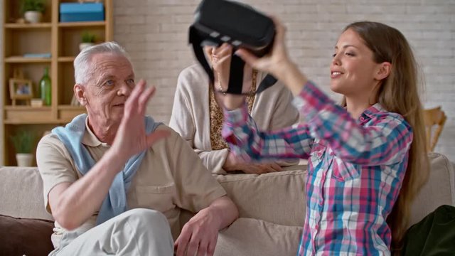 Young woman in VR headset sitting beside elderly man, then putting it on him and explaining how it works